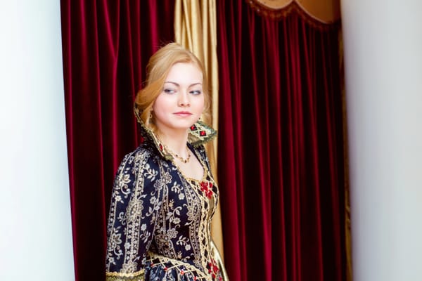 A blond woman in an ornate gown with a high collar against velvet curtains.